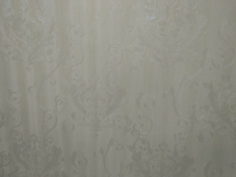 A close-up view of an intricate wallpaper pattern featuring ornate, swirling designs with floral and leaf motifs in a subtle, embossed texture. The background is a soft, muted beige color, giving it an elegant and classic appearance.