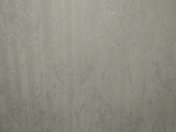 A close-up view of an intricate wallpaper pattern featuring ornate, swirling designs with floral and leaf motifs in a subtle, embossed texture. The background is a soft, muted beige color, giving it an elegant and classic appearance.