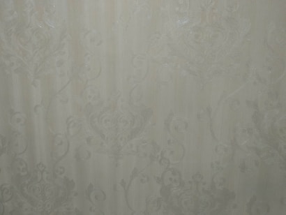 A close-up view of an intricate wallpaper pattern featuring ornate, swirling designs with floral and leaf motifs in a subtle, embossed texture. The background is a soft, muted beige color, giving it an elegant and classic appearance.