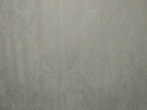 A close-up view of an intricate wallpaper pattern featuring ornate, swirling designs with floral and leaf motifs in a subtle, embossed texture. The background is a soft, muted beige color, giving it an elegant and classic appearance.