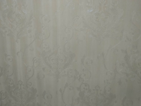 A close-up view of an intricate wallpaper pattern featuring ornate, swirling designs with floral and leaf motifs in a subtle, embossed texture. The background is a soft, muted beige color, giving it an elegant and classic appearance.