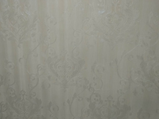 A close-up view of an intricate wallpaper pattern featuring ornate, swirling designs with floral and leaf motifs in a subtle, embossed texture. The background is a soft, muted beige color, giving it an elegant and classic appearance.