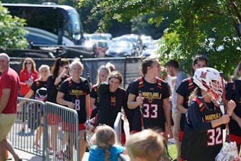 A group of young women wearing Maryland sports uniforms walk together outdoors, carrying lacrosse sticks and helmets. They appear to be a sports team, possibly arriving or leaving a game. There are people in the background, including spectators and others walking by. Foliage is visible to the right, with a tree providing shade.