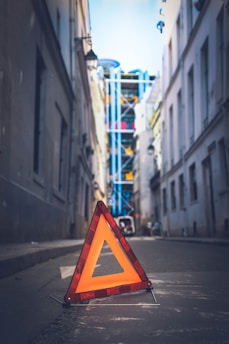 A bright orange triangular warning sign is placed on a narrow street between tall buildings. In the background, a modern structure with colorful, vertical pipes is visible, suggesting an urban setting.