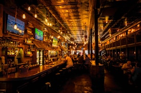A dimly lit bar interior with a warm ambiance, featuring several television screens displaying sports broadcasts above a wooden counter. Patrons are seated at the bar and tables, engaged in conversation. The ceiling is ornate with decorative panels and hanging light fixtures add a cozy glow to the scene.
