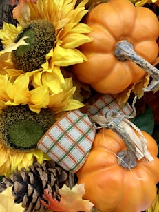 Sunflowers with bright yellow petals are arranged next to small, round orange pumpkins. A plaid ribbon with green and orange stripes adds a decorative touch. Pinecones and autumn leaves surround the arrangement, enhancing the fall theme.