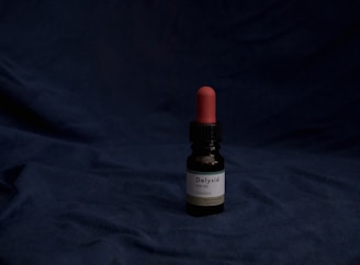 A small dark brown glass bottle with a black dropper cap and a red rubber top is placed on a dark blue fabric background. The label on the bottle reads 'Delysid (LSD 25) Sandoz'.