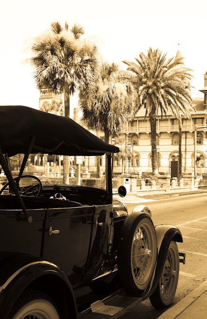 A vintage car, likely from the early 20th century, is parked on a street. The vehicle appears to be in good condition with its distinct rounded wheels and dark-colored body. In the background, there are tall palm trees and a historic building with ornate architecture, including arches and spire-like structures, suggesting a warm climate and possibly a tourist or coastal area.