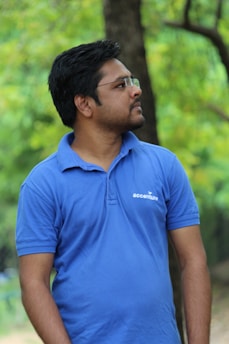 A person wearing a blue polo shirt with white writing on it stands against a natural backdrop of green foliage and trees. The person is looking to the side, and the image is slightly blurred in the background, focusing on the subject.