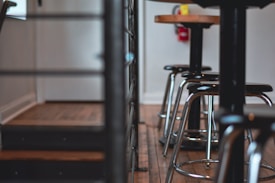 A row of metal bar stools with black seats and chrome bases is aligned along a wooden floor. The stools are accompanied by tall, black tables with wooden tops. In the background, a fire extinguisher is mounted on a light-colored wall.