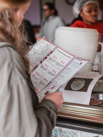 A person with blonde hair, wearing a green jacket, is holding a menu with detailed options labeled TV 1 to TV 7. In the background, a person wearing a hairnet and red shirt is seen near a stack of white plates. The scene appears to take place in a casual restaurant setting.