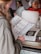 A person with blonde hair, wearing a green jacket, is holding a menu with detailed options labeled TV 1 to TV 7. In the background, a person wearing a hairnet and red shirt is seen near a stack of white plates. The scene appears to take place in a casual restaurant setting.