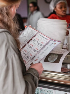 A person with blonde hair, wearing a green jacket, is holding a menu with detailed options labeled TV 1 to TV 7. In the background, a person wearing a hairnet and red shirt is seen near a stack of white plates. The scene appears to take place in a casual restaurant setting.