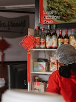 A display of various Asian sauces and condiments is arranged on a shelf, with bottles featuring labels in different languages. Above, a sign offers options of beef, chicken, and pork. A person wearing red clothing and a hairnet is partially visible in the foreground. A decorative red knot hangs from the shelf, adding a traditional touch.
