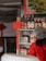 A display of various Asian sauces and condiments is arranged on a shelf, with bottles featuring labels in different languages. Above, a sign offers options of beef, chicken, and pork. A person wearing red clothing and a hairnet is partially visible in the foreground. A decorative red knot hangs from the shelf, adding a traditional touch.