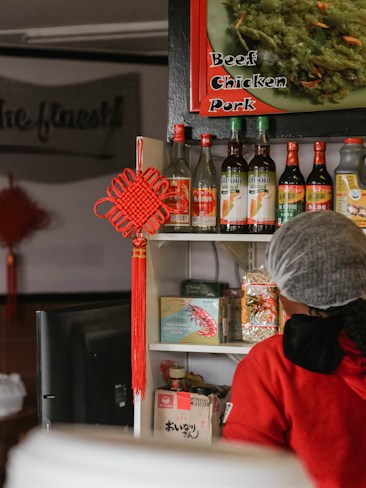 A display of various Asian sauces and condiments is arranged on a shelf, with bottles featuring labels in different languages. Above, a sign offers options of beef, chicken, and pork. A person wearing red clothing and a hairnet is partially visible in the foreground. A decorative red knot hangs from the shelf, adding a traditional touch.