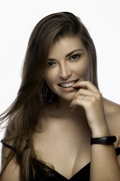 A woman with long brown hair is smiling gently, with one hand near her mouth. She is wearing a black spaghetti strap top and a black bracelet on her wrist. The lighting is soft, highlighting her natural features against a plain white background.