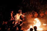 A group of people is gathered in a circular formation, watching a central figure dressed in an elaborate costume with a mask and headdress, performing a dance around an open fire. The scene is illuminated by the fire's glow, revealing the audience seated in the background.