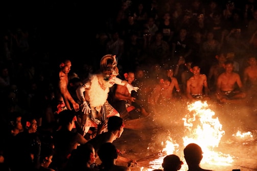 A group of people is gathered in a circular formation, watching a central figure dressed in an elaborate costume with a mask and headdress, performing a dance around an open fire. The scene is illuminated by the fire's glow, revealing the audience seated in the background.