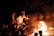 A group of people is gathered in a circular formation, watching a central figure dressed in an elaborate costume with a mask and headdress, performing a dance around an open fire. The scene is illuminated by the fire's glow, revealing the audience seated in the background.