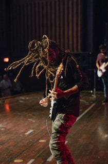 A musician with long dreadlocks energetically plays an electric guitar on stage. The atmosphere appears lively and intense, with other band members blurred in the background. The stage is dimly lit.