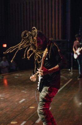 A musician with long dreadlocks energetically plays an electric guitar on stage. The atmosphere appears lively and intense, with other band members blurred in the background. The stage is dimly lit.
