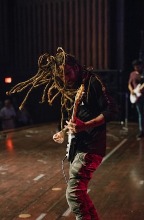 A musician with long dreadlocks energetically plays an electric guitar on stage. The atmosphere appears lively and intense, with other band members blurred in the background. The stage is dimly lit.