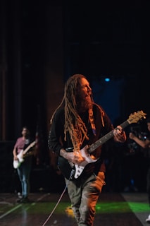 A musician with long dreadlocks passionately plays an electric guitar on stage. The background is dimly lit, with another guitarist in the distance and some stage equipment visible.