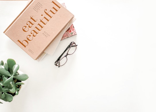 A neatly arranged flat lay features a book titled 'eat beautiful' with a beige cover, a pair of stylish eyeglasses with dark frames, and a small green succulent plant in a white pot. The items are placed on a clean, white surface, creating an organized and aesthetically pleasing composition.