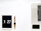 A minimalistic arrangement of digital devices placed on a white surface. On the left, there is a tablet displaying the time 1:27, next to it is a smartwatch with a brown strap. On the right, a laptop with visible keyboard and touchpad, accompanied by a smartphone. The setup suggests a modern, technological workspace.