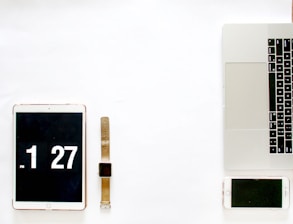 A minimalistic arrangement of digital devices placed on a white surface. On the left, there is a tablet displaying the time 1:27, next to it is a smartwatch with a brown strap. On the right, a laptop with visible keyboard and touchpad, accompanied by a smartphone. The setup suggests a modern, technological workspace.