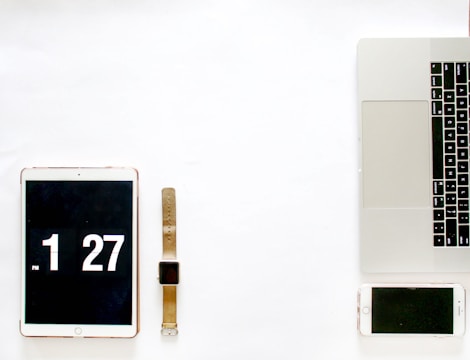 A minimalistic arrangement of digital devices placed on a white surface. On the left, there is a tablet displaying the time 1:27, next to it is a smartwatch with a brown strap. On the right, a laptop with visible keyboard and touchpad, accompanied by a smartphone. The setup suggests a modern, technological workspace.