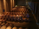 A dimly lit wine cellar filled with wooden barrels stacked neatly in rows. The room features stone walls and suspended lighting fixtures. A metal ladder is placed among the barrels, indicating an area where workers may access elevated rows.
