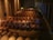 A dimly lit wine cellar filled with wooden barrels stacked neatly in rows. The room features stone walls and suspended lighting fixtures. A metal ladder is placed among the barrels, indicating an area where workers may access elevated rows.