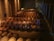 A dimly lit wine cellar filled with wooden barrels stacked neatly in rows. The room features stone walls and suspended lighting fixtures. A metal ladder is placed among the barrels, indicating an area where workers may access elevated rows.