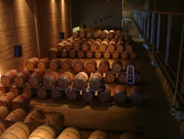 A dimly lit wine cellar filled with wooden barrels stacked neatly in rows. The room features stone walls and suspended lighting fixtures. A metal ladder is placed among the barrels, indicating an area where workers may access elevated rows.