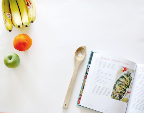 A kitchen scene featuring a wooden spoon placed beside an open cookbook with a recipe for avocado salad with fresh herbs and pepitas. On the left, there's a bunch of bananas, a green apple, and an orange arranged on a clean, white surface.