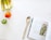 A kitchen scene featuring a wooden spoon placed beside an open cookbook with a recipe for avocado salad with fresh herbs and pepitas. On the left, there's a bunch of bananas, a green apple, and an orange arranged on a clean, white surface.