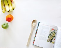 A kitchen scene featuring a wooden spoon placed beside an open cookbook with a recipe for avocado salad with fresh herbs and pepitas. On the left, there's a bunch of bananas, a green apple, and an orange arranged on a clean, white surface.
