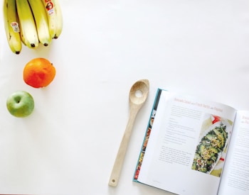 A kitchen scene featuring a wooden spoon placed beside an open cookbook with a recipe for avocado salad with fresh herbs and pepitas. On the left, there's a bunch of bananas, a green apple, and an orange arranged on a clean, white surface.