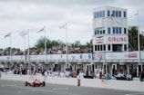 A vintage car is racing on a track in front of a crowded grandstand building. Spectators are gathered on multiple levels under covered stands, observing the race. The grandstand features prominent signage and branding, including a tower with large clocks and flags. People are dressed in vintage attire, enhancing the classic motorsport ambiance.