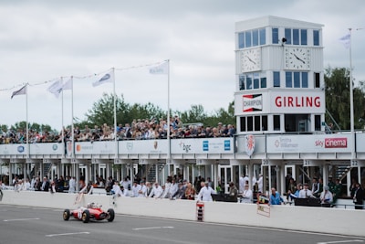 A vintage car is racing on a track in front of a crowded grandstand building. Spectators are gathered on multiple levels under covered stands, observing the race. The grandstand features prominent signage and branding, including a tower with large clocks and flags. People are dressed in vintage attire, enhancing the classic motorsport ambiance.
