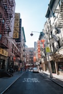 Buildings with a mix of English and Chinese signage line a narrow street. Several pedestrians walk along the sidewalks, and a car is parked in the street, which has the word 'SCHOOL' painted on it. Fire escapes are visible on the buildings, and the atmosphere suggests a busy, urban environment.