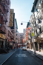 Buildings with a mix of English and Chinese signage line a narrow street. Several pedestrians walk along the sidewalks, and a car is parked in the street, which has the word 'SCHOOL' painted on it. Fire escapes are visible on the buildings, and the atmosphere suggests a busy, urban environment.