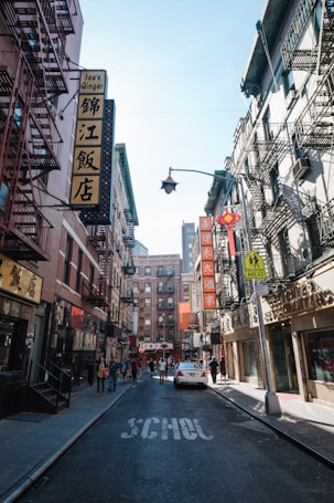 Buildings with a mix of English and Chinese signage line a narrow street. Several pedestrians walk along the sidewalks, and a car is parked in the street, which has the word 'SCHOOL' painted on it. Fire escapes are visible on the buildings, and the atmosphere suggests a busy, urban environment.