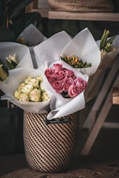 Several bouquets of flowers wrapped in white paper are arranged inside a decorative woven basket. The flowers include pale yellow and pink roses, as well as lilies and other blooms. The background features soft shadows and natural textures, creating a cozy and inviting atmosphere.