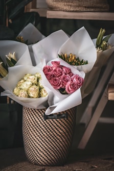 Several bouquets of flowers wrapped in white paper are arranged inside a decorative woven basket. The flowers include pale yellow and pink roses, as well as lilies and other blooms. The background features soft shadows and natural textures, creating a cozy and inviting atmosphere.