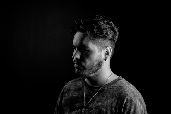 A black and white portrait of a person with short, styled hair and a beard. The person is wearing a T-shirt and necklace, looking contemplatively to the side, with a neutral expression. The lighting creates a dramatic contrast, highlighting the contours of the face against a dark background.
