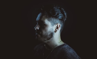 A profile view of a man with short, styled hair and a beard against a dark background. He is wearing a gold chain and a small earring. The lighting highlights the contours of his face, creating a dramatic shadow.
