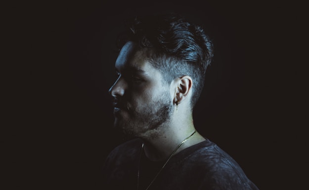 A profile view of a man with short, styled hair and a beard against a dark background. He is wearing a gold chain and a small earring. The lighting highlights the contours of his face, creating a dramatic shadow.